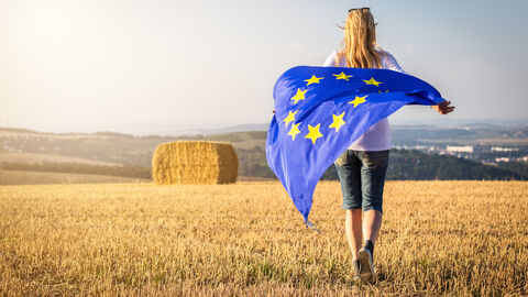 Eine Frau läuft mit wehender Flagge der Europäischen Union über eine landwirtschaftliche Fläche. Eine Frau läuft mit wehender Flagge der Europäischen Union über eine landwirtschaftliche Fläche.