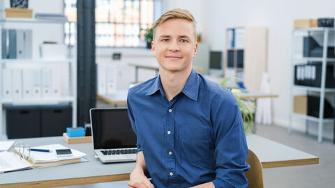 Ein junger Mann sitzt am Schreibtisch und lächelt in die Kamera.