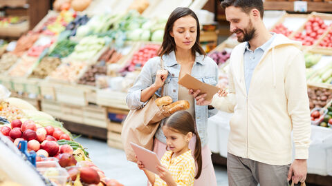 Eine junge Familie steht vor einem Obststand und blickt auf den Einkaufszettel.