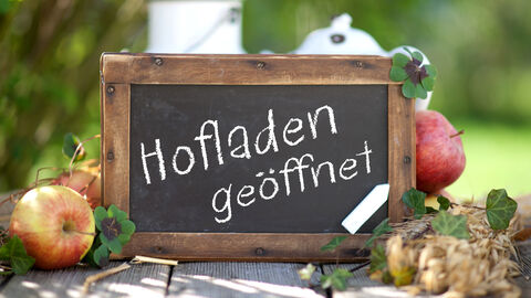Eine Tafel mit der Aufschrift "Hofladen geöffnet" steht zwischen Äpfeln in der Natur.