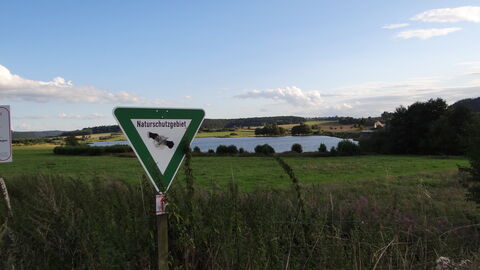 Ein Naturschutzgebiets-Schild steht vor einer Wiesen- und Seenfläche. Ein Naturschutzgebiets-Schild steht vor einer Wiesen- und Seenfläche.