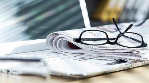 Eine Brille liegt zusammen mit Tageszeitungen vor einem Notebook.