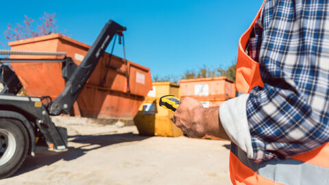 Ein Mann in Warnweste verlädt einen Bauschuttcontainer mit einer Fernbedienung auf einen LKW.