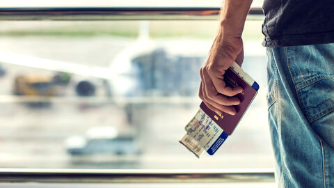 Ein Mann hält ein Flugticket und den Reisepass in der Hand während im Hintergrund ein Flugzeug parkt. Ein Mann hält ein Flugticket und den Reisepass in der Hand während im Hintergrund ein Flugzeug parkt.
