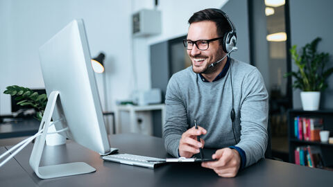Ein Mann mit Headset sitzt lächelnd am Schreibtisch und blickt auf seinen Monitor. Ein Mann mit Headset sitzt lächelnd am Schreibtisch und blickt auf seinen Monitor.