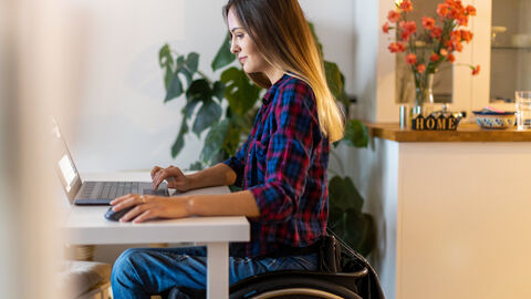 Eine junge Frau sitzt im Rollstuhl und arbeitet am Notebook.