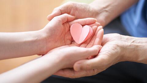 Hände von einer jungen und einer alten Person halten zusammen ein Papierherz.