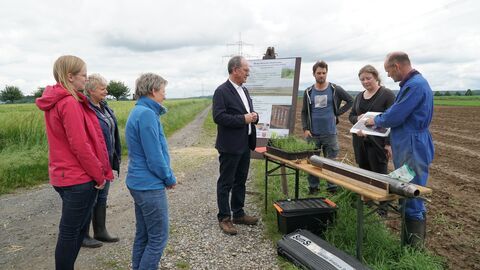 Besuch in Ahlbach Ein Mann und sechs weitere Personen stehen auf einem Feld, der Mann informiert die anderen über Miscanthus.
