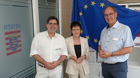 Regierungspräsident Dr. Christoph Ullrich begrüßt die Landesvorsitzende der Europa-Union, Ute Wiegand-Fleischhacker, zum Austausch mit Michael Schär, dem Leiter des EU-Informationszentrums. Sie stehen nebeneinander vor einer Europaflagge.